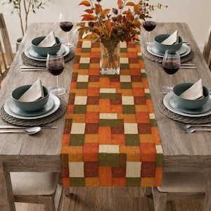 K&ouml;nnte beinhalten: Ein rechteckiger Tischl&auml;ufer mit einem Patchwork-Muster aus orangefarbenen, braunen, olivgr&uuml;nen und cremefarbenen Quadraten. Der L&auml;ufer liegt mittig auf einem h&ouml;lzernen Esstisch mit vier Gedecken, die jeweils eine Schale, einen Teller und ein Weinglas umfassen.