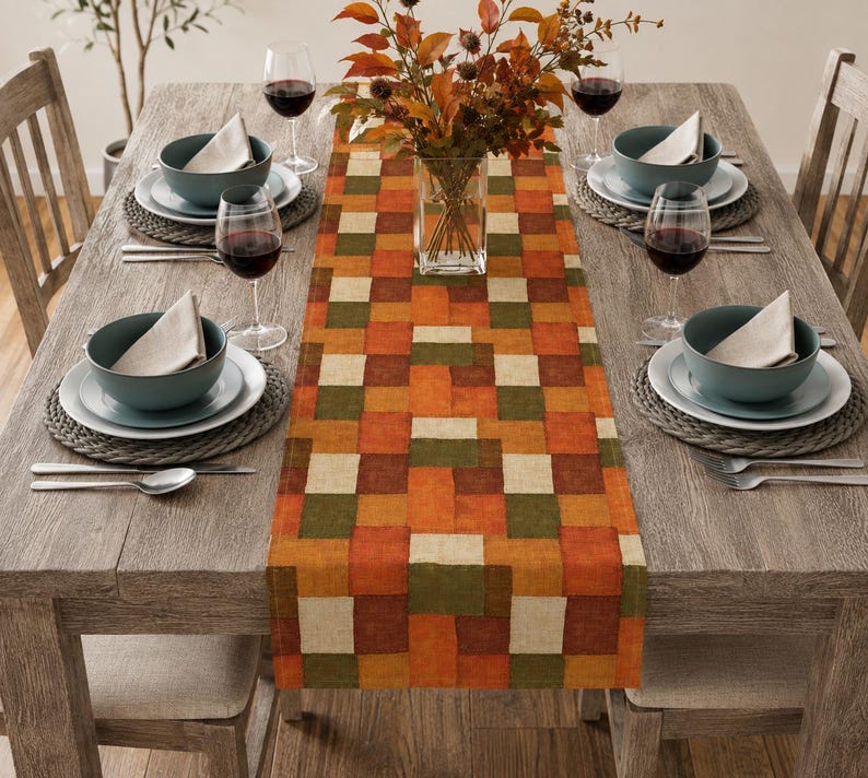 K&ouml;nnte beinhalten: Ein rechteckiger Tischl&auml;ufer mit einem Patchwork-Muster aus orangefarbenen, braunen, olivgr&uuml;nen und cremefarbenen Quadraten. Der L&auml;ufer liegt mittig auf einem h&ouml;lzernen Esstisch mit vier Gedecken, die jeweils eine Schale, einen Teller und ein Weinglas umfassen.