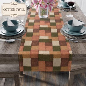 K&ouml;nnte beinhalten: Ein Tischl&auml;ufer aus Baumwollk&ouml;per mit einem geometrischen Patchwork-Muster in Olivgr&uuml;n, Terrakotta, Braun und Creme. Der L&auml;ufer liegt mittig auf einem Holztisch mit Gedecken, bestehend aus Sch&uuml;sseln, Tellern und Besteck.