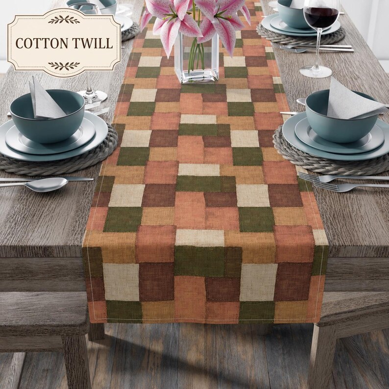 K&ouml;nnte beinhalten: Ein Tischl&auml;ufer aus Baumwollk&ouml;per mit einem geometrischen Patchwork-Muster in Olivgr&uuml;n, Terrakotta, Braun und Creme. Der L&auml;ufer liegt mittig auf einem Holztisch mit Gedecken, bestehend aus Sch&uuml;sseln, Tellern und Besteck.