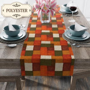 K&ouml;nnte beinhalten: Ein Tischl&auml;ufer mit geometrischem Blockmuster in Orange, Dunkelgr&uuml;n und Creme. Der L&auml;ufer liegt auf einem Holztisch mit Gedecken, bestehend aus blauen Schalen, wei&szlig;en Servietten und einer Glasvase mit rosa Lilien.