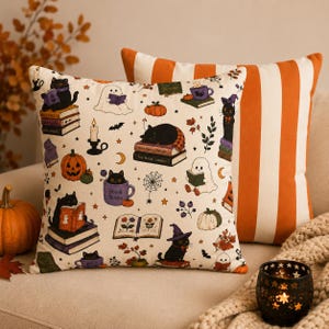 May include: A square throw pillow featuring a cream-coloured fabric with a repeating pattern of black cats, ghosts, pumpkins, books, and candles. A second pillow behind it displays vertical orange and white stripes. The print includes the text Boo & Books.