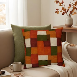 May include: A square throw pillow featuring a patchwork design of rectangular blocks in shades of burnt orange, deep burgundy, forest green, and cream. The pillow is positioned in front of a solid olive green throw pillow on a neutral sofa.