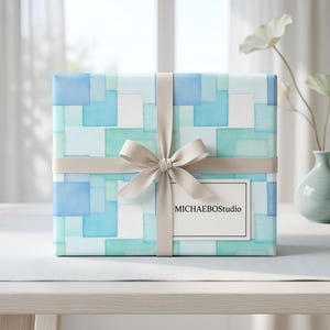 May include: A wrapped gift box with a light blue, teal, and white geometric pattern. A beige ribbon is tied around the box with a bow. A label on the front reads "MICHAELBOStudio". The gift box is on a white table.