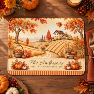 May include: A rectangular placemat featuring an autumn landscape with a farmhouse, rolling fields, and trees with orange and yellow leaves. Two baskets filled with pumpkins sit on the sides. The text reads The Andersons GRATEFUL TOGETHER.
