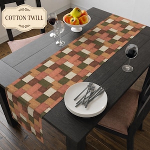 K&ouml;nnte beinhalten: Ein Tischl&auml;ufer aus Baumwollk&ouml;per mit einem geometrischen Patchwork-Muster in Rost-, Olivgr&uuml;n-, Creme- und Braunt&ouml;nen. Der L&auml;ufer liegt auf einem dunklen Holztisch mit einem Stapel wei&szlig;er Teller, Besteck und einer Schale mit &Auml;pfeln.