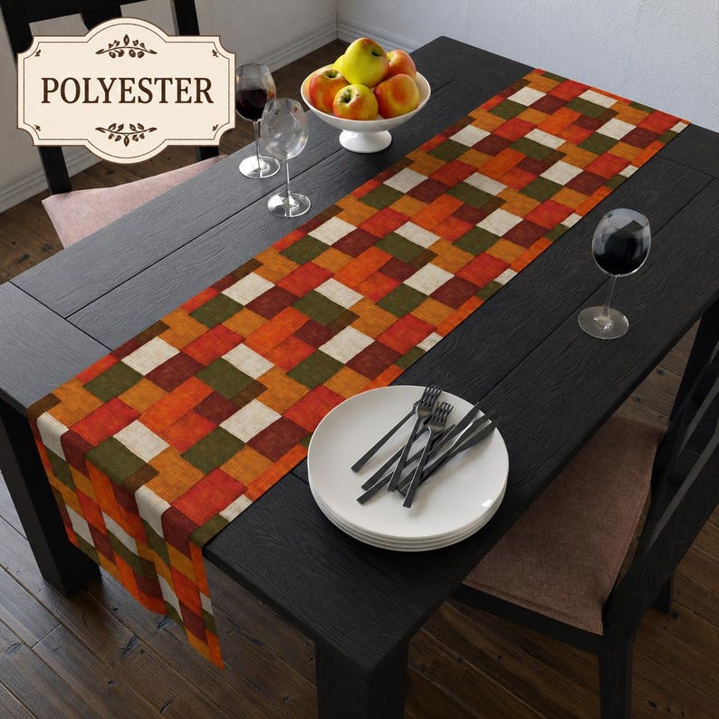 K&ouml;nnte beinhalten: Ein rechteckiger Tischl&auml;ufer mit einem geometrischen Patchwork-Muster aus Quadraten in Orange-, Dunkelgr&uuml;n- und Cremet&ouml;nen. Der Stoff ist als Polyester gekennzeichnet. Der L&auml;ufer liegt auf einem dunklen Holztisch mit Tellern und Gl&auml;sern.