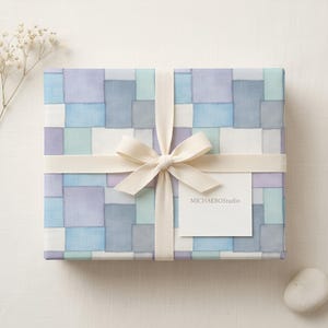 Puede incluir: Un regalo envuelto en papel con un diseño de patchwork en tonos pastel de azul, morado y blanco. Está atado con una cinta y un lazo color crema. Una pequeña etiqueta blanca con "MICHAELBOstudio" está adjunta.
