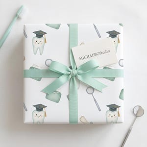 Puede incluir: Un regalo envuelto sobre un fondo blanco, con un patrón de dientes sonrientes con gorros de graduación, instrumentos dentales y recipientes de hilo dental. El regalo está atado con una cinta y un lazo verde claro. Se adjunta una pequeña tarjeta con el texto "MICHAELBOStudio".