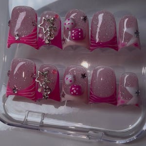 May include: A set of ten artificial nails in a clear plastic case. The nails feature a pink and glitter design with a pink base, white accents, and silver star and gem embellishments. Some nails have dice-shaped decorations.