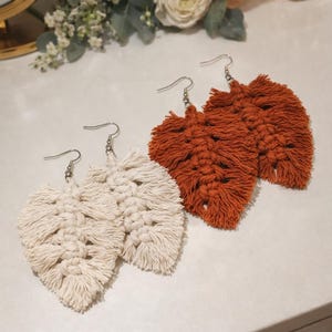 May include: Two pairs of leaf-shaped macrame earrings. One pair is off-white, the other is a burnt orange colour. The earrings have silver-coloured ear wires. The earrings are handmade and have a bohemian style.