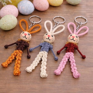 Puede incluir: Tres llaveros de conejito hechos a mano con detalles en naranja, azul y rosa. Cada conejito tiene una cabeza de madera con una cara pintada, un cuerpo de ganchillo y patas colgantes. Huevos de Pascua y flores en el fondo.