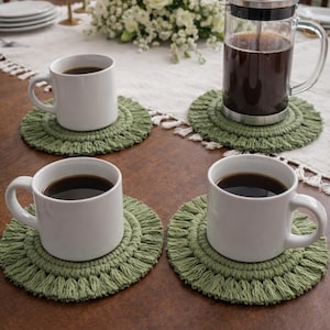 Puede incluir: Una mesa con cuatro tazas de café blancas llenas de café oscuro, sobre posavasos redondos de macramé verde. Una cafetera de émbolo y velas también están sobre la mesa, con un arreglo floral en el fondo. La mesa está puesta para una reunión.