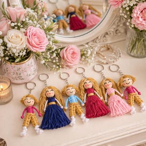 May include: A collection of handmade doll keychains with blonde yarn hair and smiling faces. The dolls wear dresses in various colors, including blue, pink, and red, and are attached to silver key rings. The background includes flowers and a mirror.