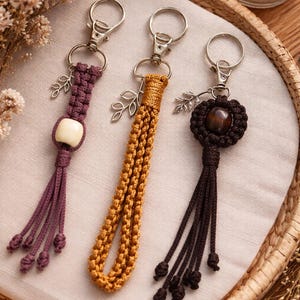 May include: Three handmade macrame keychains in shades of purple, gold, and brown. Each keychain features a metal clasp, a decorative leaf charm, and a unique macrame design with tassels. The keychains are displayed on a neutral fabric.