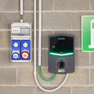 May include: An electric vehicle charging station with a dark gray casing and a green light. The station is mounted on a gray brick wall. A white and green cable is connected to the charging station. A sign with a green background is visible.