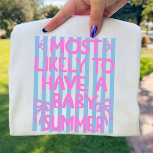 Op de afbeelding: Crèmekleurige sweatshirt met de tekst "MOST LIKELY TO HAVE A BABY SUMMER" in roze, tegen een achtergrond van lichtblauwe verticale strepen en roze strikaccenten. De sweatshirt wordt door een hand omhoog gehouden.