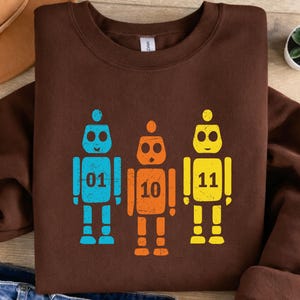 K&ouml;nnte beinhalten: Ein braunes Sweatshirt mit drei bunten Roboter-Grafiken. Die Roboter sind blau, orange und gelb, jeder mit einer anderen Nummer: 01, 10 und 11. Das Design ist auf einem weichen, bequemen Sweatshirt gedruckt.