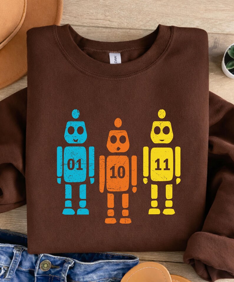 K&ouml;nnte beinhalten: Ein braunes Sweatshirt mit drei bunten Roboter-Grafiken. Die Roboter sind blau, orange und gelb, jeder mit einer anderen Nummer: 01, 10 und 11. Das Design ist auf einem weichen, bequemen Sweatshirt gedruckt.