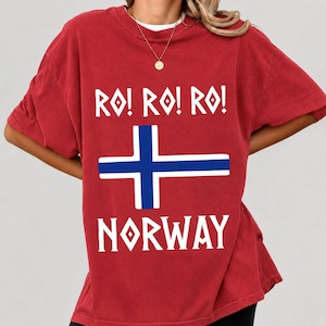 Pode incluir: Uma t-shirt vermelha de manga curta com o texto branco RO! RO! RO! acima de um gráfico de cruz azul e branca, com o texto branco NORWAY abaixo do gráfico. A t-shirt tem gola redonda e um corte descontraído, usada com um colar de corrente dourada.