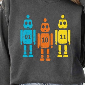 K&ouml;nnte beinhalten: Dunkelgraues Sweatshirt mit drei bunten Roboter-Grafiken. Die Roboter sind blau, orange und gelb, mit den Zahlen 01, 10 und 11. Das Design hat einen Used-Look.