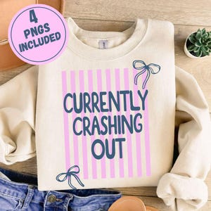 以下が含まれることがあります： クリーム色のスウェットシャツ。テキスト「CURRENTLY CRASHING OUT」は濃い青色で、ピンクの縦縞模様の上に配置されています。デザインには濃い青色のリボンアクセントが含まれています。「4 PNGS INCLUDED」と書かれたピンク色の円が左上にあります。