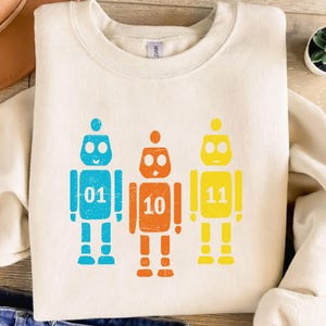 K&ouml;nnte beinhalten: Cremefarbenes Sweatshirt mit drei bunten Roboter-Grafiken. Jeder Roboter hat eine andere Farbe: blau, orange und gelb. Jeder Roboter hat eine Nummer auf der Brust: 01, 10 und 11. Die Roboter haben einen Used-Look.