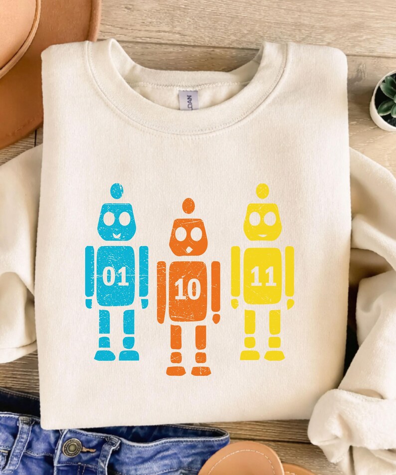 K&ouml;nnte beinhalten: Cremefarbenes Sweatshirt mit drei bunten Roboter-Grafiken. Jeder Roboter hat eine andere Farbe: blau, orange und gelb. Jeder Roboter hat eine Nummer auf der Brust: 01, 10 und 11. Die Roboter haben einen Used-Look.