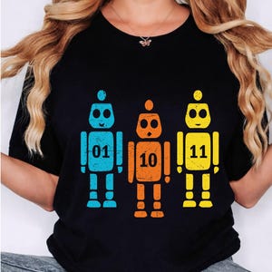 May include: Black t-shirt featuring three colorful robot illustrations. The robots are blue, orange, and yellow, each with a different number: 01, 10, and 11. The design is simple and graphic.