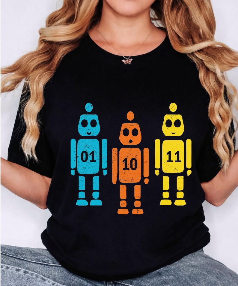 K&ouml;nnte beinhalten: Schwarzes T-Shirt mit drei bunten Roboter-Illustrationen. Die Roboter sind blau, orange und gelb, mit den Zahlen 01, 10 und 11. Das Design ist einfach und grafisch.