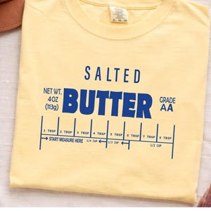 May include: A pale yellow t-shirt featuring the words "SALTED BUTTER" in blue. Below is a measuring guide with tablespoon and cup measurements. The shirt also includes the text "NET WT. 4OZ (113g) GRADE AA".