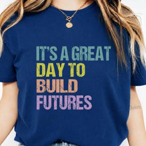 Op de afbeelding: Marineblauw T-shirt met de tekst "IT'S A GREAT DAY TO BUILD FUTURES" in teal, geel, perzik en paars. Het shirt heeft een ronde hals en korte mouwen.