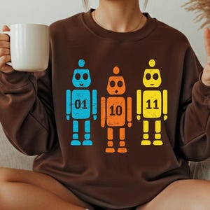K&ouml;nnte beinhalten: Ein braunes Sweatshirt mit drei bunten Roboter-Illustrationen. Die Roboter sind blau, orange und gelb, jeder mit einer Nummer: 01, 10 und 11. Der Stil ist l&auml;ssig.