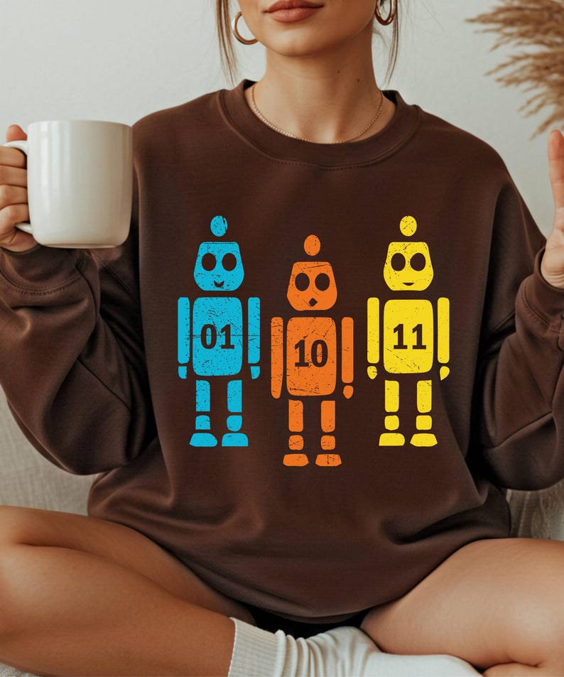 K&ouml;nnte beinhalten: Ein braunes Sweatshirt mit drei bunten Roboter-Illustrationen. Die Roboter sind blau, orange und gelb, jeder mit einer Nummer: 01, 10 und 11. Der Stil ist l&auml;ssig.