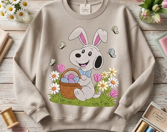 Puede incluir: Sudadera beige con un personaje de dibujos animados de Snoopy vestido de conejito de Pascua, sosteniendo una cesta de huevos coloridos. El diseño incluye mariposas y flores. La sudadera es de estilo casual y cómodo.