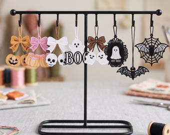 Może przedstawiać: Czarny metalowy stojak w kształcie litery T z ośmioma wiszącymi ozdobami na Halloween. Ozdoby obejmują haftowane kokardy, dynie, czaszki, duszki, ramkę z napisem HEY BOO, nietoperza oraz pajęczynę z nietoperzem.