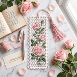 Puede incluir: Un marcapáginas rectangular blanco con un patrón de panal y un diseño bordado de rosas rosas con hojas y tallos verdes. El marcapáginas incluye una borla de seda rosa unida a la parte superior y está rodeado de rosas rosas.