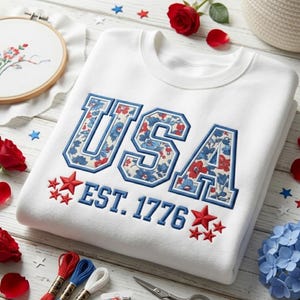 May include: A white crewneck sweatshirt featuring the embroidered text USA in a block font filled with a red and blue floral pattern. Below the text, the words EST. 1776 are embroidered in blue, flanked by small red embroidered stars.