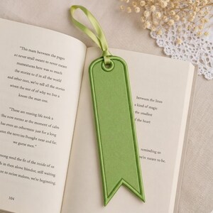 May include: A green bookmark with a ribbon loop, resting on the open pages of a book. The bookmark has a rectangular shape with a pointed bottom and a stitched border. The ribbon matches the bookmark's color.