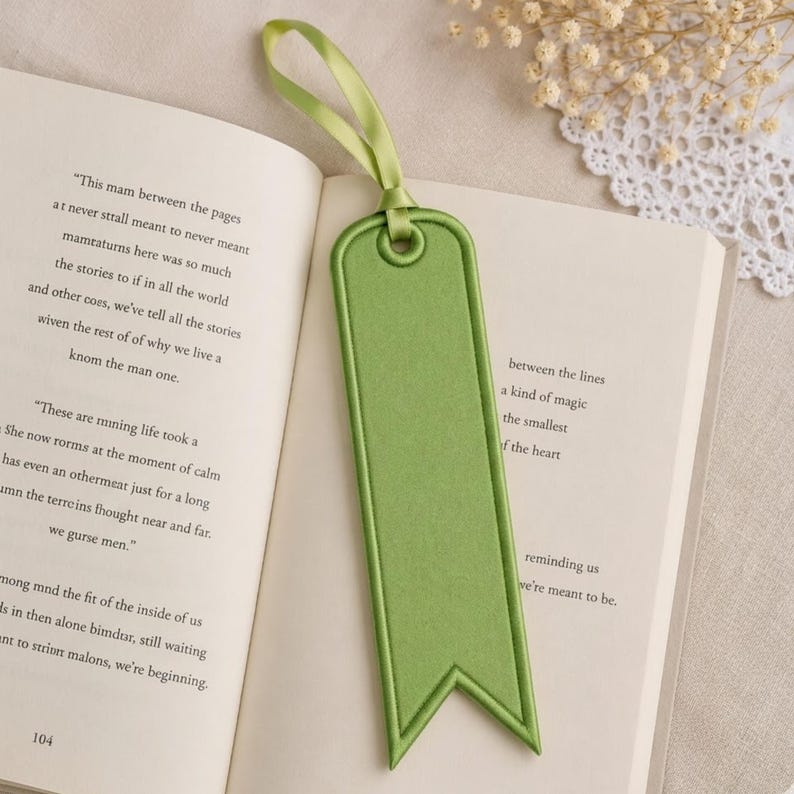 May include: A green bookmark with a ribbon loop, resting on the open pages of a book. The bookmark has a rectangular shape with a pointed bottom and a stitched border. The ribbon matches the bookmark's color.