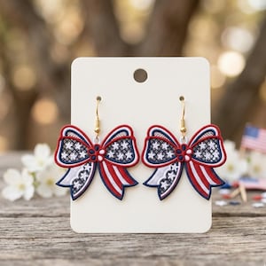May include: A pair of dangling earrings featuring embroidered bow shapes with a red, white, and blue color scheme. The design includes white stars on a blue background and red and white stripes, finished with gold-toned metal earring hooks.