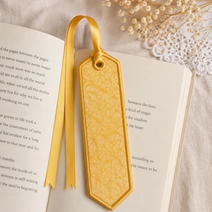 May include: A yellow bookmark with a decorative pattern and a matching ribbon is placed in an open book. The bookmark has a stitched border and a textured surface. The book's pages are visible, with text in black.