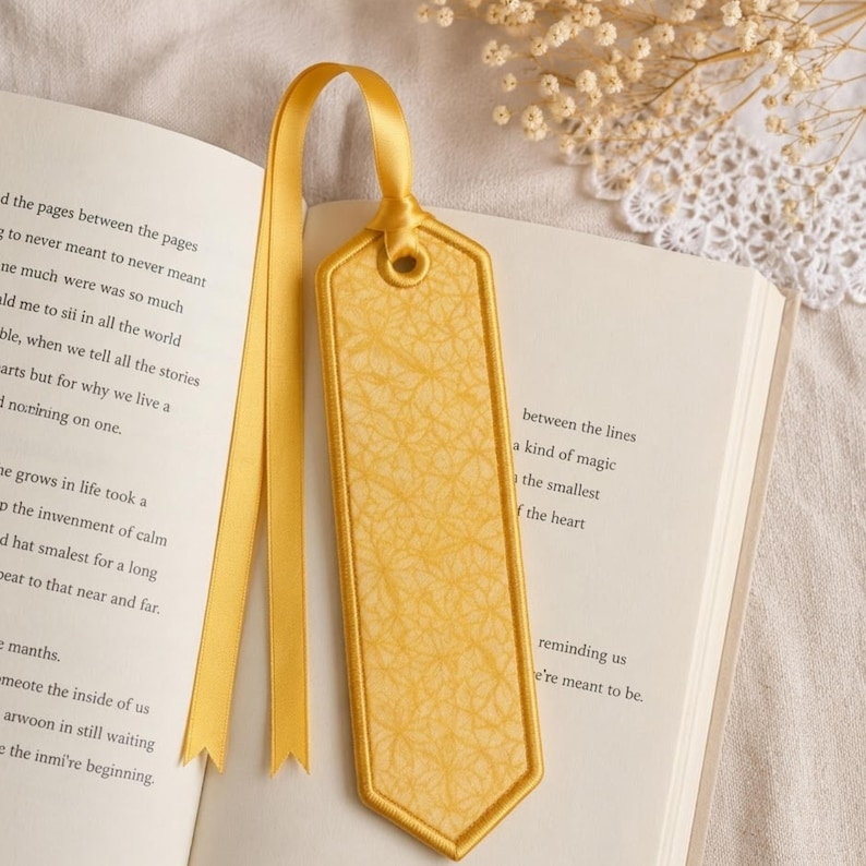 May include: A yellow bookmark with a decorative pattern and a matching ribbon is placed in an open book. The bookmark has a stitched border and a textured surface. The book's pages are visible, with text in black.