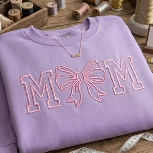 May include: A lilac-colored sweatshirt with the word "MOM" embroidered in pink, featuring a bow design between the letters. A gold necklace with the word "mom" is also visible. The sweatshirt is on a wooden surface.