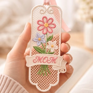 Op de afbeelding: Een witte bloemenbladwijzer met een roze lint. De bladwijzer heeft een boeket bloemen in roze, paars en wit, met het woord "MOM" op een banner. De bladwijzer wordt in een hand gehouden.