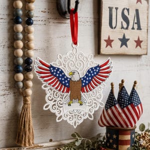 Puede incluir: Un adorno bordado blanco con un &aacute;guila calva con las alas extendidas, que incorpora los colores de la bandera estadounidense. El adorno cuelga de una cinta roja. Un letrero de madera con "USA" y estrellas est&aacute; en el fondo.