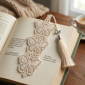 May include: An off-white lace bookmark featuring three butterfly motifs and small floral accents. The bookmark is attached to a thin cord with a small silver-toned dragonfly charm, a white bead, and a long, matching off-white fabric tassel.