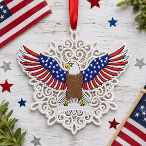 Puede incluir: Adorno calado blanco con un &aacute;guila calva bordada con las alas extendidas. Las alas muestran las estrellas y las rayas de la bandera estadounidense. Se adjunta una cinta roja para colgarlo. Estrellas decorativas y banderas americanas en el fondo.