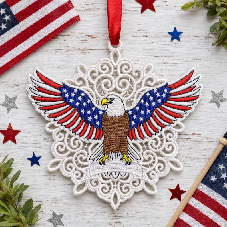 Puede incluir: Adorno calado blanco con un &aacute;guila calva bordada con las alas extendidas. Las alas muestran las estrellas y las rayas de la bandera estadounidense. Se adjunta una cinta roja para colgarlo. Estrellas decorativas y banderas americanas en el fondo.