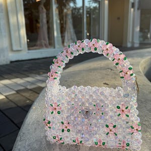 Könnte beinhalten: Eine transparente, perlenbesetzte Handtasche mit Henkel und floralen Akzenten. Die Tasche ist mit rosa und grünen Blumendetails verziert. Der Henkel ist ebenfalls mit rosa und grünen Blumen verziert. Die Tasche liegt auf einer Steinoberfläche.